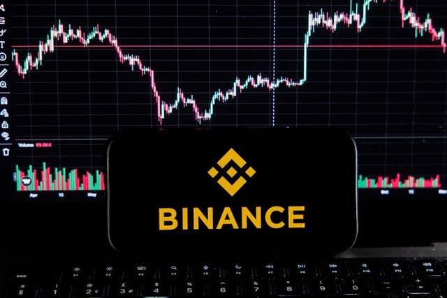 Logotipo da Binance em tela de celular diante de gráficos de criptomoedas